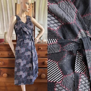May include: A sleeveless wrap dress with a geometric pattern in shades of gray, white, and red. The dress features a V-neckline, a matching belt, and a knee-length hemline. The fabric has a textured appearance.