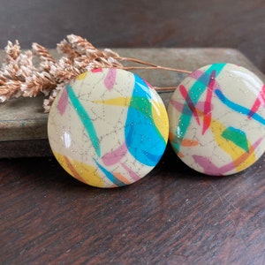 Può includere: Un paio di orecchini rotondi bianchi con un design astratto colorato. Gli orecchini hanno uno sfondo bianco con schizzi di blu, verde, giallo, rosa e rosso.