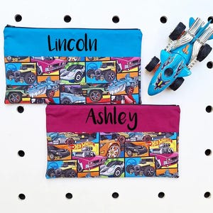 May include: Two personalised pencil cases with a Hot Wheels car print. The top case is blue with the name "Lincoln" embroidered on it. The bottom case is burgundy with the name "Ashley" embroidered on it.