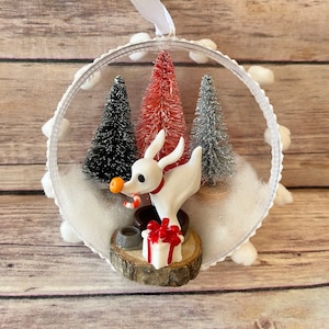 Può includere: Un ornamento di vetro trasparente con una statuetta di cane bianco e peloso all'interno. Il cane tiene un bastoncino di zucchero e una piccola palla arancione. Ci sono tre alberi di Natale in miniatura all'interno dell'ornamento, uno rosso, uno nero e uno argentato. L'ornamento è decorato con pompon bianchi attorno al bordo.