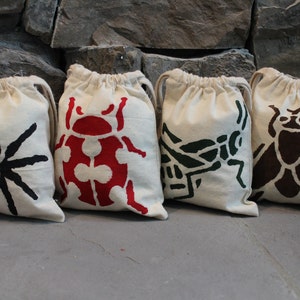 Pode incluir: Quatro pequenas bolsas de tecido branco com cordão de ajuste e diferentes desenhos de insetos. As bolsas apresentam uma aranha preta, uma joaninha vermelha, um gafanhoto verde e uma mosca marrom.