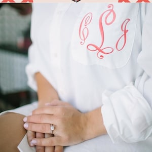 May include: A white button-down shirt with a pink embroidered monogram "S.C.J." on the chest. The shirt is worn by a person with a diamond ring on their finger.