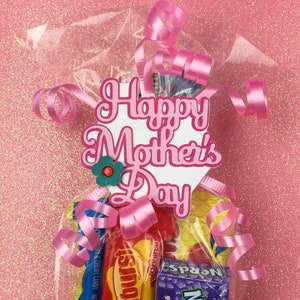 May include: A cellophane bag filled with sweets, tied with pink ribbon and a tag saying "Happy Mother's Day". The tag has a pink and white design with a small flower. The bag contains various branded sweets.