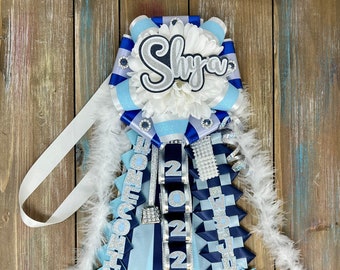May include: A blue and white graduation sash with silver glitter, a white flower, and the name "Shyla" on it. The sash also has the year "2022" and the words "Senior Class" on it. There is a blue wolf head with white claws on the bottom of the sash.