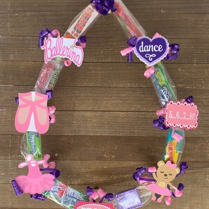 May include: A circular candy wreath featuring various wrapped sweets, purple curling ribbons, and decorative cardstock cutouts. The cutouts include a pink ballet slipper, a pink tutu, a bear in a tutu, and signs reading Ballerina, dance, 5...6...7...8!, and REGAN.