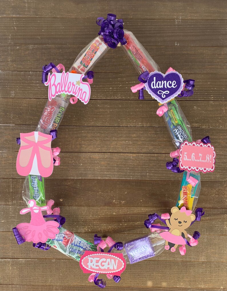 May include: A circular candy wreath featuring various wrapped sweets, purple curling ribbons, and decorative cardstock cutouts. The cutouts include a pink ballet slipper, a pink tutu, a bear in a tutu, and signs reading Ballerina, dance, 5...6...7...8!, and REGAN.
