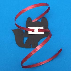 May include: A black fox-shaped cutout with a white rectangle attached, and a red ribbon spiraling around it. The fox has a tail and is set against a blue background.