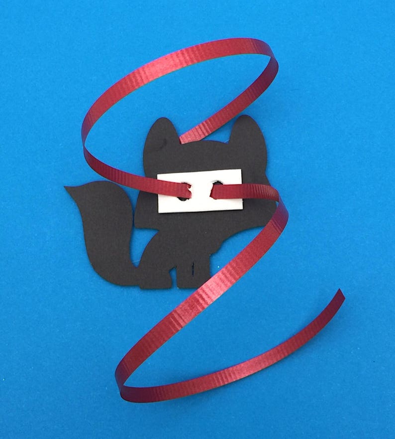 May include: A black fox-shaped cutout with a white rectangle attached, and a red ribbon spiraling around it. The fox has a tail and is set against a blue background.