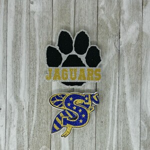 K&ouml;nnte beinhalten: Zwei glitzernde B&uuml;gelbilder. Das erste B&uuml;gelbild ist ein schwarzer Pfotenabdruck mit dem Wort "JAGUARS" in goldenen Glitzerbuchstaben auf silbernem Hintergrund. Das zweite B&uuml;gelbild ist ein blauer und goldener Glitzerbuchstabe "S" mit Sternen und Streifen.