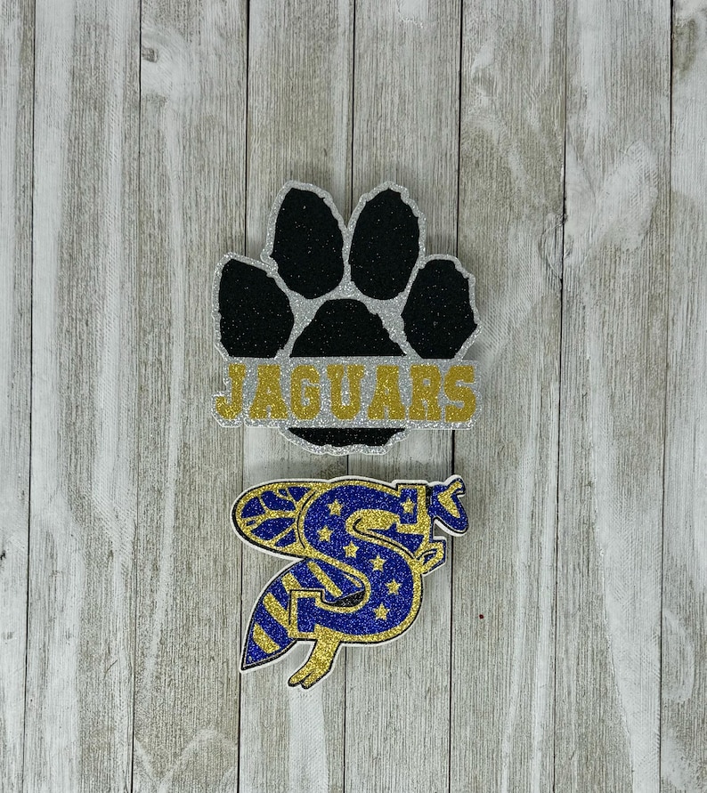 K&ouml;nnte beinhalten: Zwei glitzernde B&uuml;gelbilder. Das erste B&uuml;gelbild ist ein schwarzer Pfotenabdruck mit dem Wort "JAGUARS" in goldenen Glitzerbuchstaben auf silbernem Hintergrund. Das zweite B&uuml;gelbild ist ein blauer und goldener Glitzerbuchstabe "S" mit Sternen und Streifen.