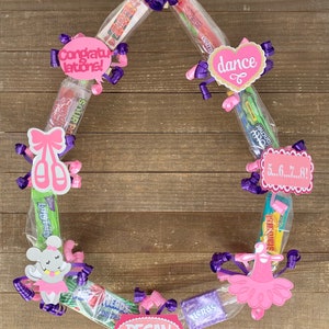 May include: A circular candy wreath constructed from various wrapped candy bars and boxes, decorated with purple curling ribbons and pink paper cutouts featuring the text Congratulations, dance, 5...6...7...8!, and the name REGAN.