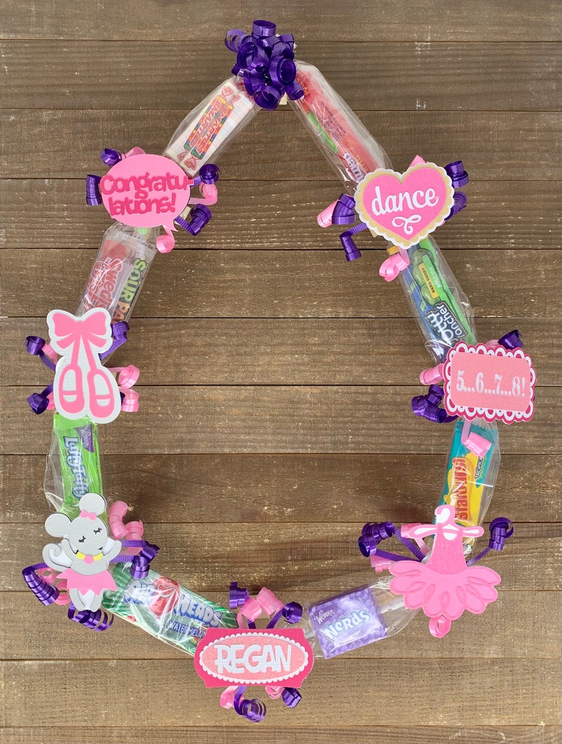 May include: A circular candy wreath constructed from various wrapped candy bars and boxes, decorated with purple curling ribbons and pink paper cutouts featuring the text Congratulations, dance, 5...6...7...8!, and the name REGAN.
