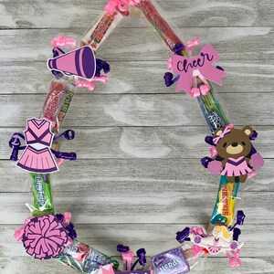 May include: A pink and purple cheerleader-themed candy wreath. The wreath is decorated with candy tubes, bows, and cutouts of a megaphone, a cheerleader uniform, a pom-pom, a bear, a cheerleader, and the word "Cheer". The name "Lacy" is also included.