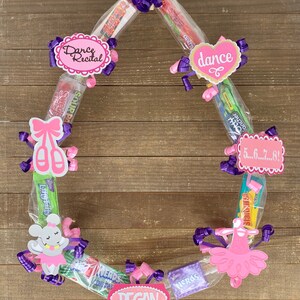 May include: A circular candy wreath featuring various boxed and bagged sweets arranged in a ring. The wreath is decorated with purple and pink curling ribbons, paper cutouts of ballet slippers, a tutu, a mouse, and signs reading Dance Recital, dance, 5...6...7...8!, and REGAN.