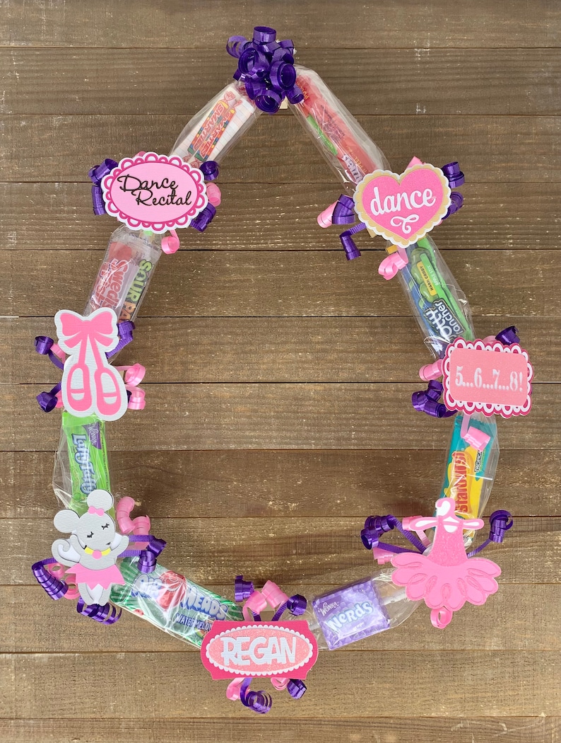 May include: A circular candy wreath featuring various boxed and bagged sweets arranged in a ring. The wreath is decorated with purple and pink curling ribbons, paper cutouts of ballet slippers, a tutu, a mouse, and signs reading Dance Recital, dance, 5...6...7...8!, and REGAN.