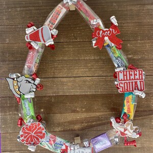 May include: A circular candy wreath featuring various wrapped sweets including Smarties, Sour Patch Kids, Jolly Ranchers, Starburst, and Nerds. The wreath is decorated with red and silver ribbons, a bulldog cutout, a megaphone, and the text Cheer Squad and Kari.