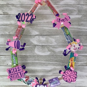 May include: A circular candy wreath featuring various snack packages, pink curling ribbons, and paper cutouts. Decorations include a purple megaphone, a cheerleader figure, a pom-pom, and signs reading 2022, Cheer, Cheer Squad, and LACY.