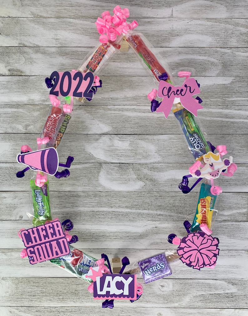 May include: A circular candy wreath featuring various snack packages, pink curling ribbons, and paper cutouts. Decorations include a purple megaphone, a cheerleader figure, a pom-pom, and signs reading 2022, Cheer, Cheer Squad, and LACY.