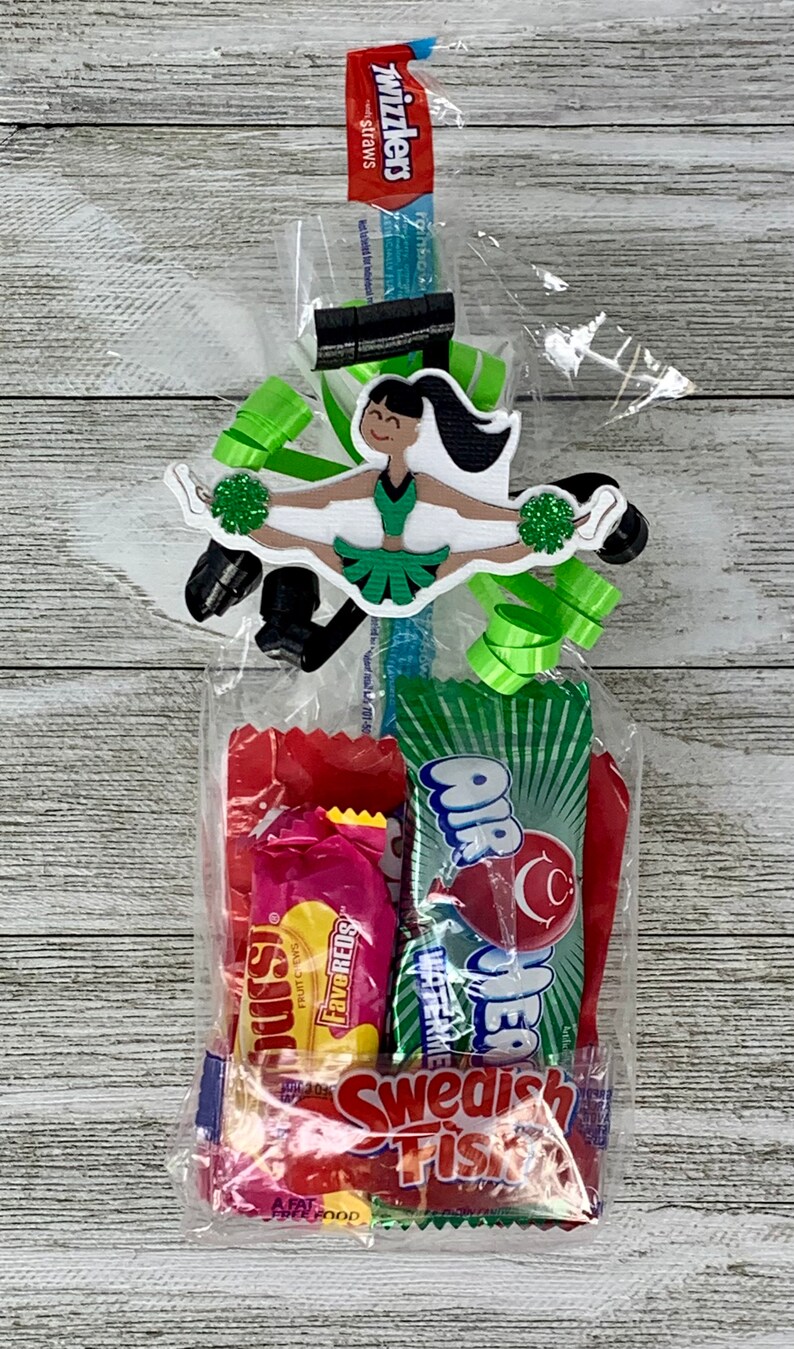May include: A clear plastic gift bag containing assorted candy including Twizzlers, AirHeads, and Swedish Fish. The bag is decorated with a die-cut paper cheerleader figure in a green and black uniform holding green pom-poms, accented with green ribbons.