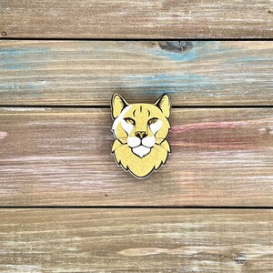 K&ouml;nnte beinhalten: Ein goldbrauner Holzausschnitt eines Gepardenkopfes mit wei&szlig;en Markierungen um die Augen und die Nase. Der Gepard schaut direkt in die Kamera.