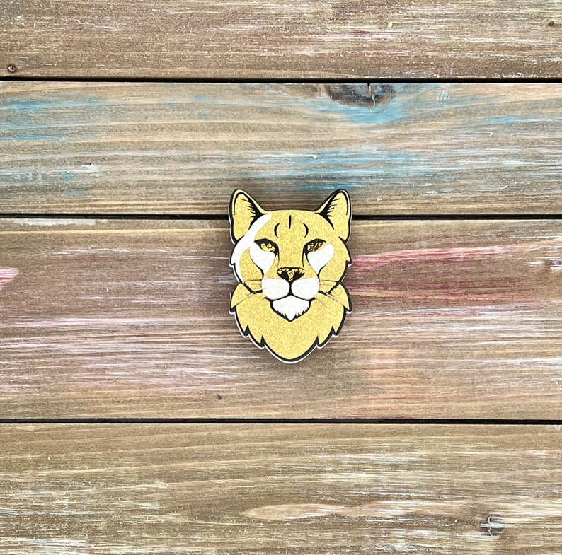K&ouml;nnte beinhalten: Ein goldbrauner Holzausschnitt eines Gepardenkopfes mit wei&szlig;en Markierungen um die Augen und die Nase. Der Gepard schaut direkt in die Kamera.