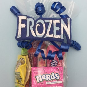 May include: A clear plastic gift bag filled with assorted sweets, including a pink box of Nerds Seriously Strawberry and a yellow packet of Extreme Sour hard candy. The bag is topped with a blue felt sign reading FROZEN and blue ribbons.