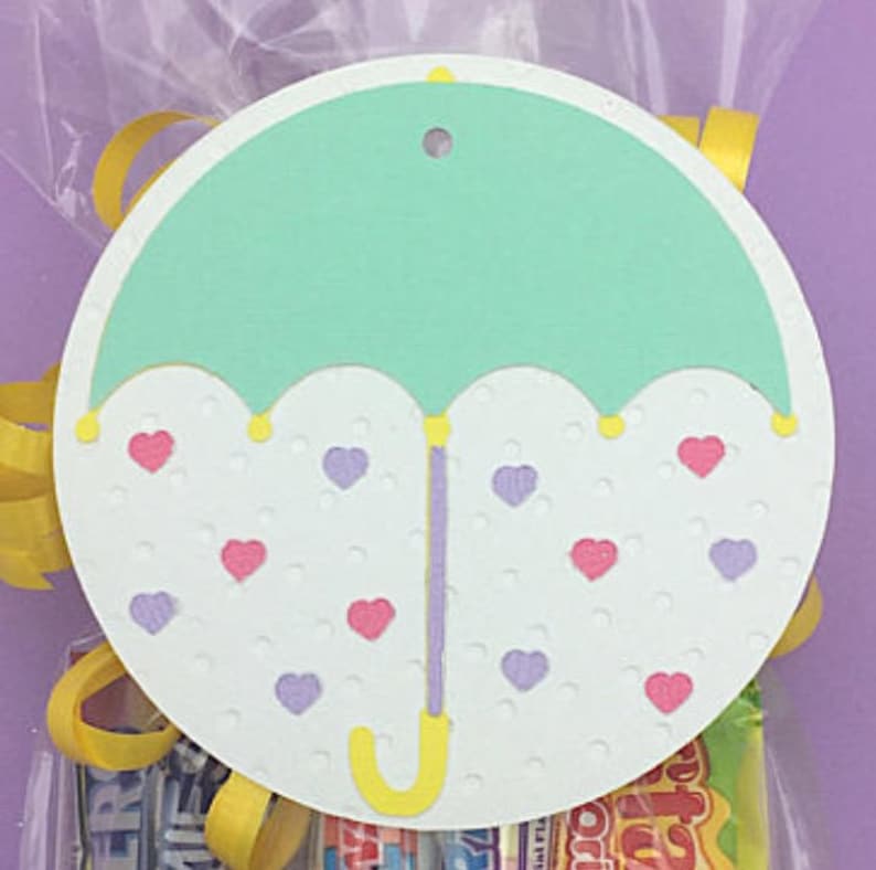 May include: A circular gift tag with a light green umbrella design, yellow handle and accents. The umbrella sits on a white background with pink and purple heart accents. A small hole is at the top for hanging. The tag is in front of a cellophane bag.