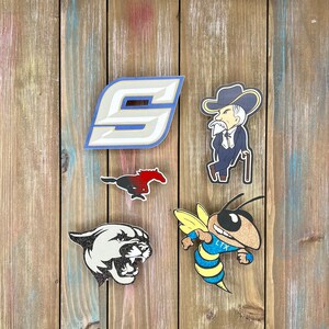 K&ouml;nnte beinhalten: F&uuml;nf verschiedene Schulmaskottchen-Logos auf einem Holzgrund. Die Logos sind eine blaue und wei&szlig;e Nummer "9", ein rotes und schwarzes Pferd, ein wei&szlig;er Pantherkopf, eine gelbe und blaue Biene mit dem Text "LTX" und ein Cartoon-Mann mit wei&szlig;em Bart, der einen blauen Anzug und einen blau-wei&szlig;en Cowboyhut tr&auml;gt.