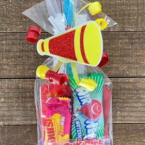 May include: A clear plastic treat bag filled with assorted candies including Starburst FaveReds, AirHeads Watermelon, and Swedish Fish. The bag is sealed with a yellow and red glittery megaphone cutout and curled red and yellow ribbons.