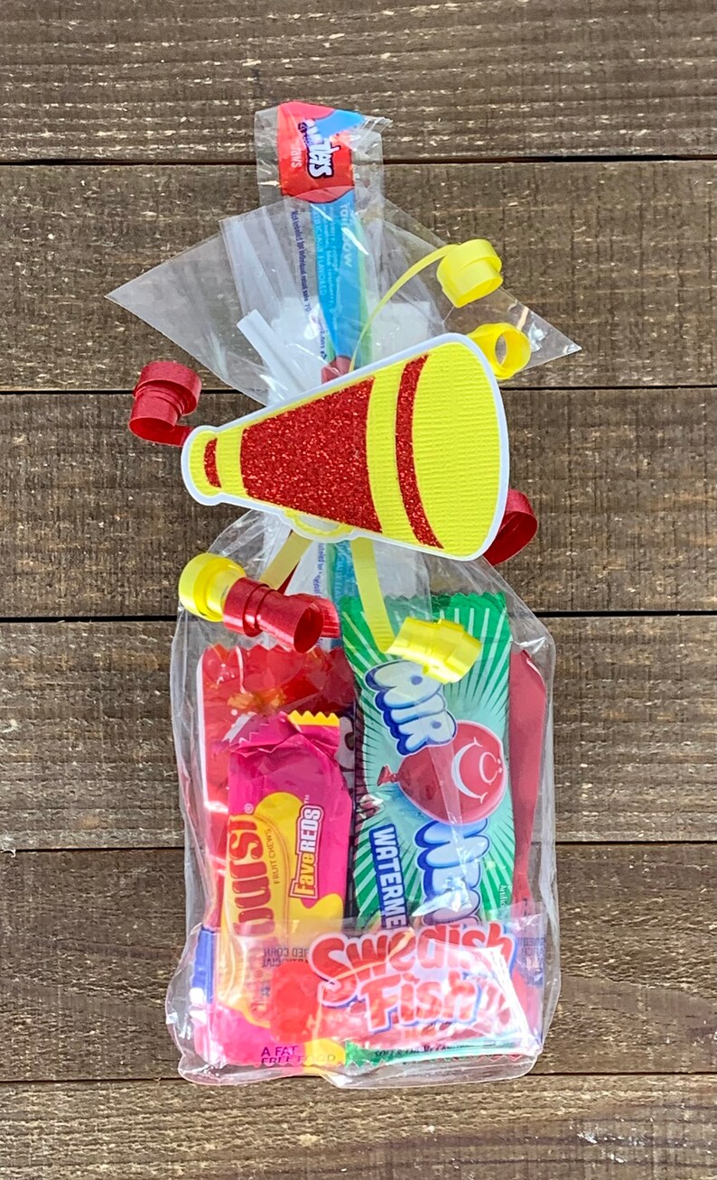 May include: A clear plastic treat bag filled with assorted candies including Starburst FaveReds, AirHeads Watermelon, and Swedish Fish. The bag is sealed with a yellow and red glittery megaphone cutout and curled red and yellow ribbons.