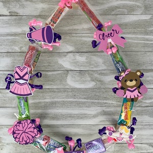 May include: A circular candy wreath constructed from various individually wrapped snacks, decorated with pink and purple cheerleading-themed cutouts, including a megaphone, a uniform, a bear, and a bow with the word Cheer, plus the name LACY.