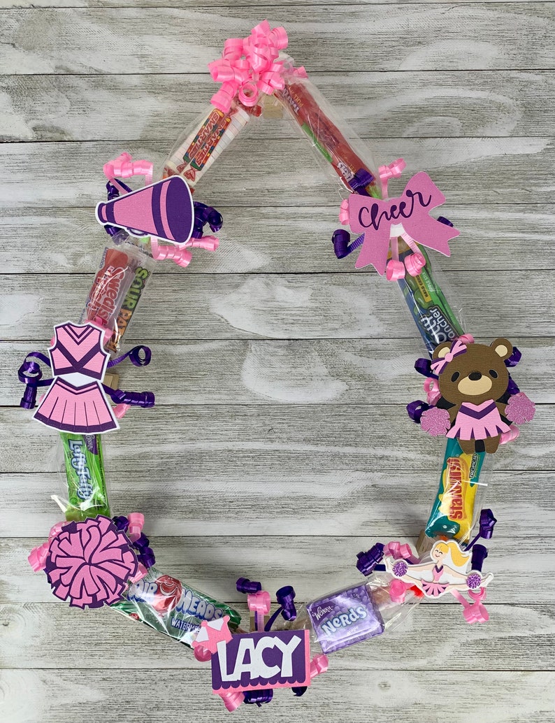 May include: A circular candy wreath constructed from various individually wrapped snacks, decorated with pink and purple cheerleading-themed cutouts, including a megaphone, a uniform, a bear, and a bow with the word Cheer, plus the name LACY.