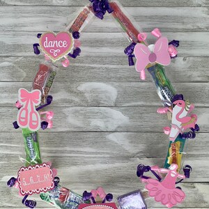 May include: A circular candy lei featuring various wrapped sweets, purple curling ribbons, and pink paper cutouts. Decorations include a heart with the word dance, ballet slippers, a flamingo, a tutu, and a sign with the name REGAN and 5...6...7...8!.