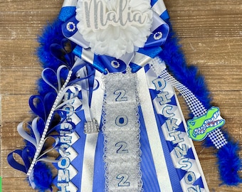 May include: A blue and white homecoming mum with the name "Malia" and the year "2022" on it. The mum also has a blue and white checkered ribbon with an alligator charm.