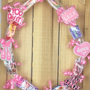 May include: A circular candy wreath decorated with pink curling ribbons, assorted candy bars, and pink paper cutouts featuring the text "YOU DID IT!", "5...6...7...8!", "dance", "Tiny Dancer", and "PAISLEY", along with a pair of pink ballet slippers.