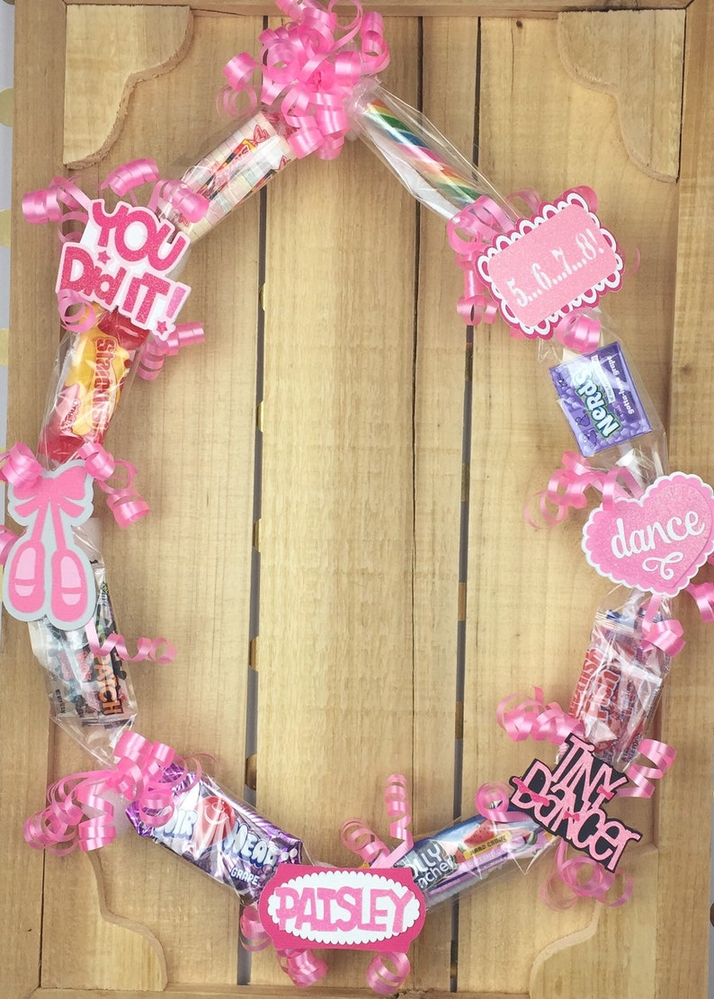 May include: A circular candy wreath decorated with pink curling ribbons, assorted candy bars, and pink paper cutouts featuring the text "YOU DID IT!", "5...6...7...8!", "dance", "Tiny Dancer", and "PAISLEY", along with a pair of pink ballet slippers.