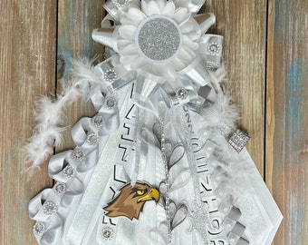 May include: A white and silver homecoming mum featuring a central flower with a silver glitter center, surrounded by layered ribbons, white feathers, rhinestone accents, and a cutout of an eagle head. The ribbons display the text SENIOR.