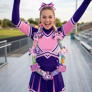 May include: A purple and pink cheerleading uniform with a matching hair bow, featuring a long-sleeved top and pleated skirt. A decorative candy lei is worn around the neck, adorned with small snack packages, a megaphone cutout, and the text 2022, LACY.