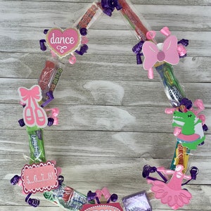 May include: A pink and purple candy lei with a dance theme. The lei includes a heart with the word "dance" on it, a ballerina shoe, a pink bow, and a green alligator. The lei also includes a pink sign with the text "Regan" and a pink sign with the text "5...6...7...8!"