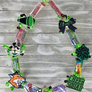 May include: A circular candy lei featuring various wrapped sweets, lime green curling ribbons, and black glittery cutouts including a megaphone, a bow with the word Cheer, a cheerleader, a pom-pom, a fox, a Cheer Squad sign, and a name tag reading Sydney.