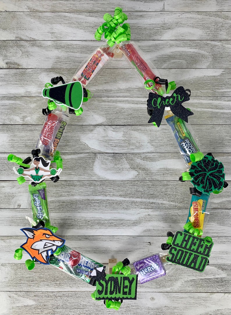 May include: A circular candy lei featuring various wrapped sweets, lime green curling ribbons, and black glittery cutouts including a megaphone, a bow with the word Cheer, a cheerleader, a pom-pom, a fox, a Cheer Squad sign, and a name tag reading Sydney.