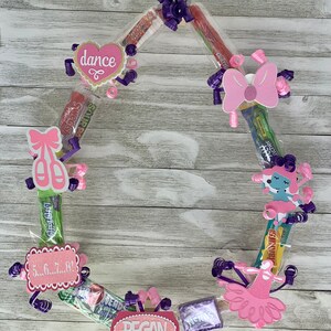 May include: A circular candy wreath constructed from various individually wrapped sweets, decorated with purple curling ribbons and pink paper cutouts featuring the text dance, 5...6...7...8!, and the name REGAN, alongside ballet-themed icons.