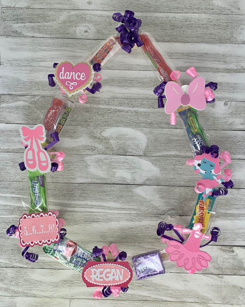 May include: A circular candy wreath constructed from various individually wrapped sweets, decorated with purple curling ribbons and pink paper cutouts featuring the text dance, 5...6...7...8!, and the name REGAN, alongside ballet-themed icons.