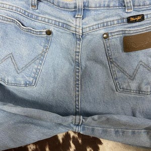 May include: Light blue denim jeans with the Wrangler logo on a brown leather patch. The jeans have two back pockets with the signature Wrangler stitching and brass rivets. The jeans are a classic style.