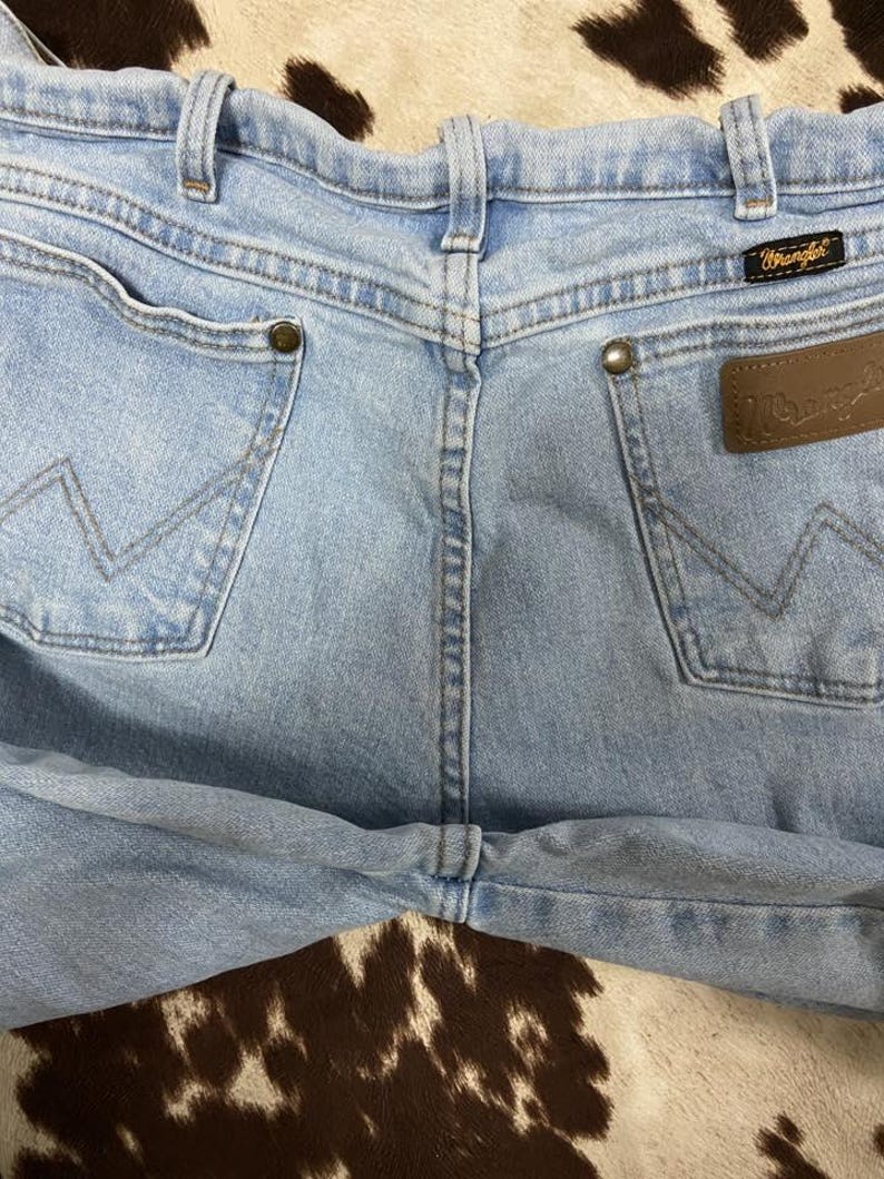 May include: Light blue denim jeans with the Wrangler logo on a brown leather patch. The jeans have two back pockets with the signature Wrangler stitching and brass rivets. The jeans are a classic style.