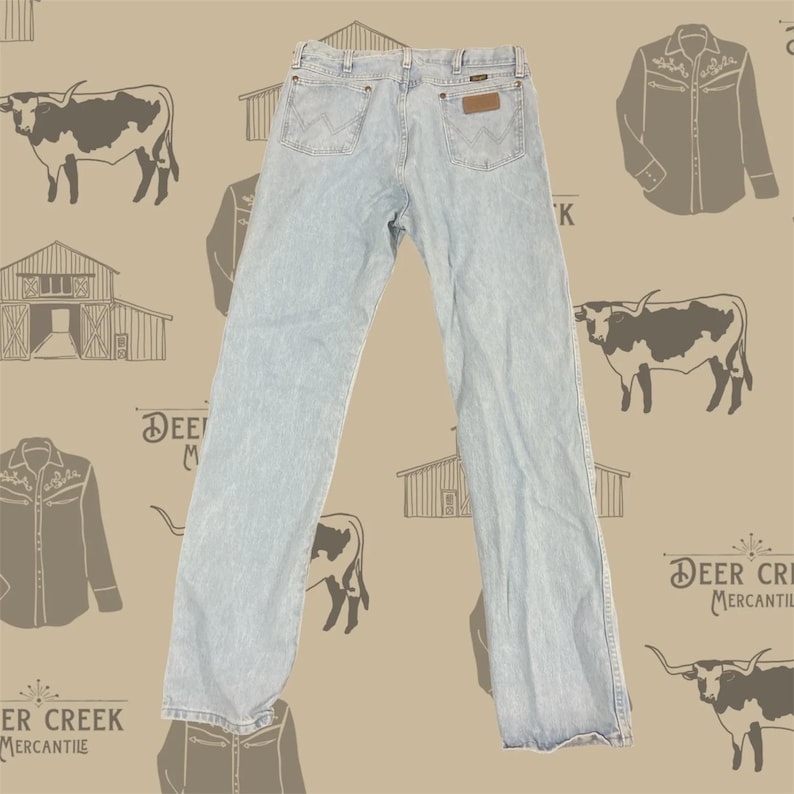 Pu&ograve; includere: Jeans in denim azzurro con un classico design a cinque tasche, chiusura con bottone e cerniera, e una toppa in pelle marrone sulla tasca posteriore destra con il logo del marchio Wrangler visibile sul retro del capo.