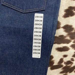 May include: Close-up of a pair of dark blue denim jeans. The jeans have a classic five-pocket design with a stitched back pocket detail. A white tag displays the size 28x38. The jeans are made of a durable denim fabric.