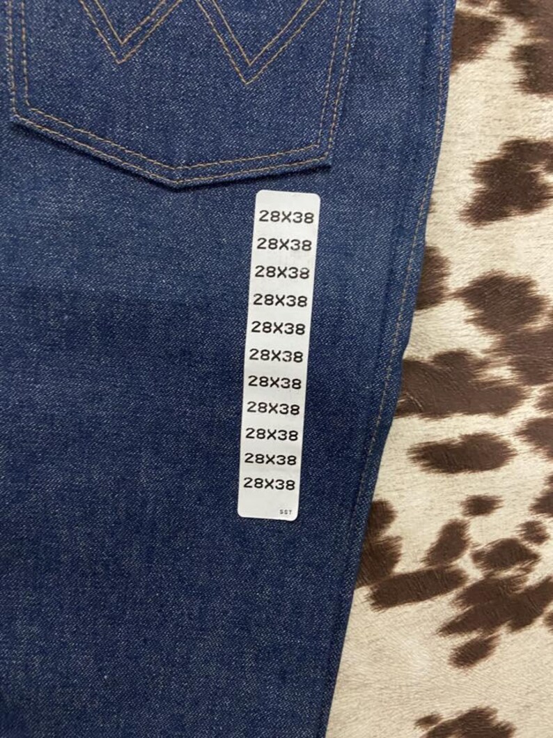 May include: Close-up of a pair of dark blue denim jeans. The jeans have a classic five-pocket design with a stitched back pocket detail. A white tag displays the size 28x38. The jeans are made of a durable denim fabric.