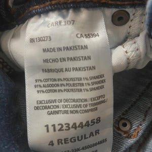May include: A white clothing care label attached to blue denim fabric. The label displays text including CARE307, RN 130273, CA 55394, Made in Pakistan, 91% cotton, 8% polyester, 1% spandex, Exclusive of decoration, 112344458, and 4 Regular.