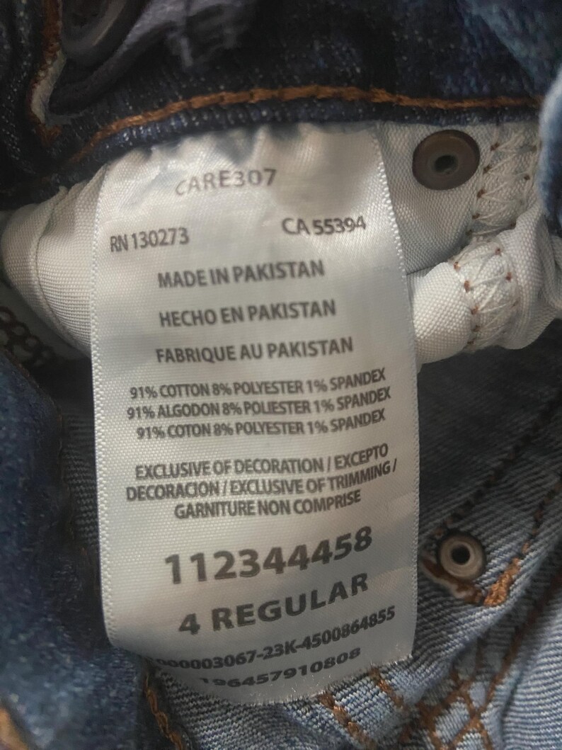 May include: A white clothing care label attached to blue denim fabric. The label displays text including CARE307, RN 130273, CA 55394, Made in Pakistan, 91% cotton, 8% polyester, 1% spandex, Exclusive of decoration, 112344458, and 4 Regular.